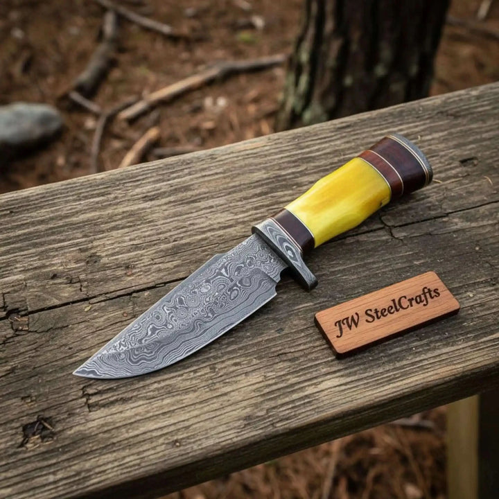 Sharp hunting knife with yellow handle, Damascus pattern blade, brown leather sheath, and outdoor wooden backdrop.