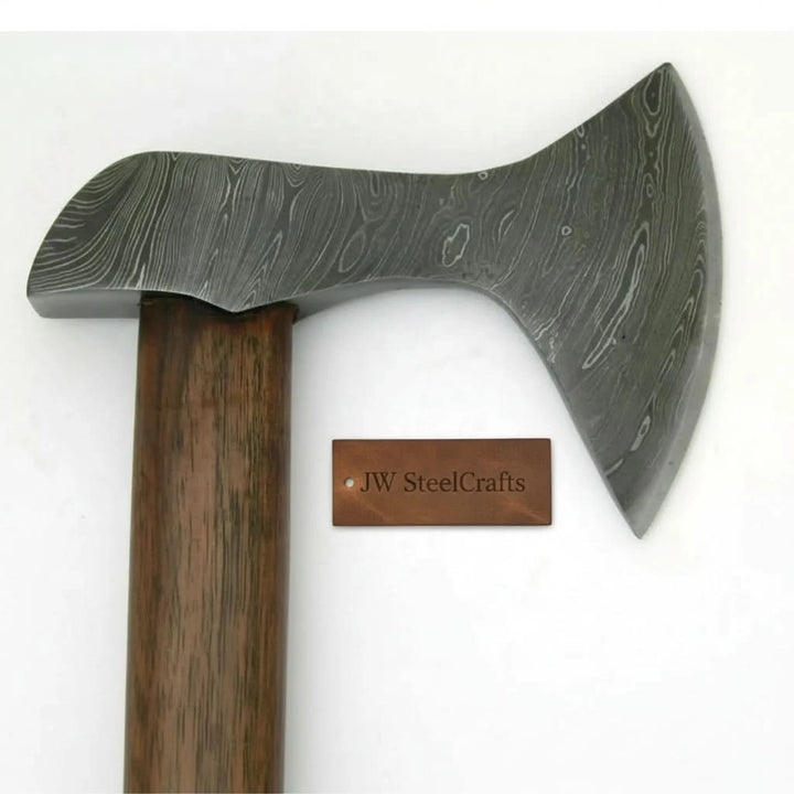     Custom bushcraft Damascus axe featuring polished steel craftsmanship and dark wooden grip displayed in woodland environment.