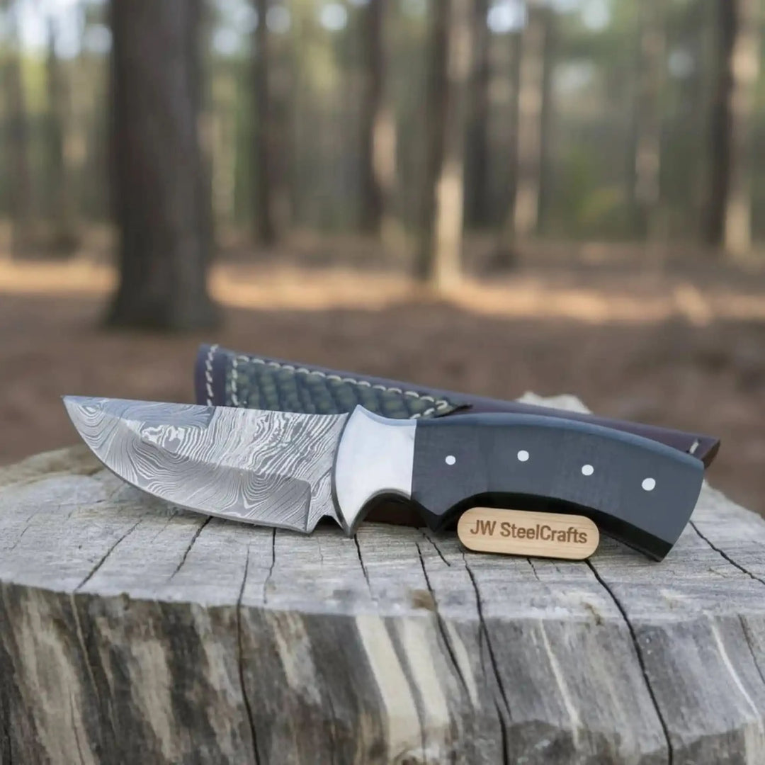 Rustic fixed blade Damascus knife with black grip, curved blade, leather sheath, and blurred forest background.