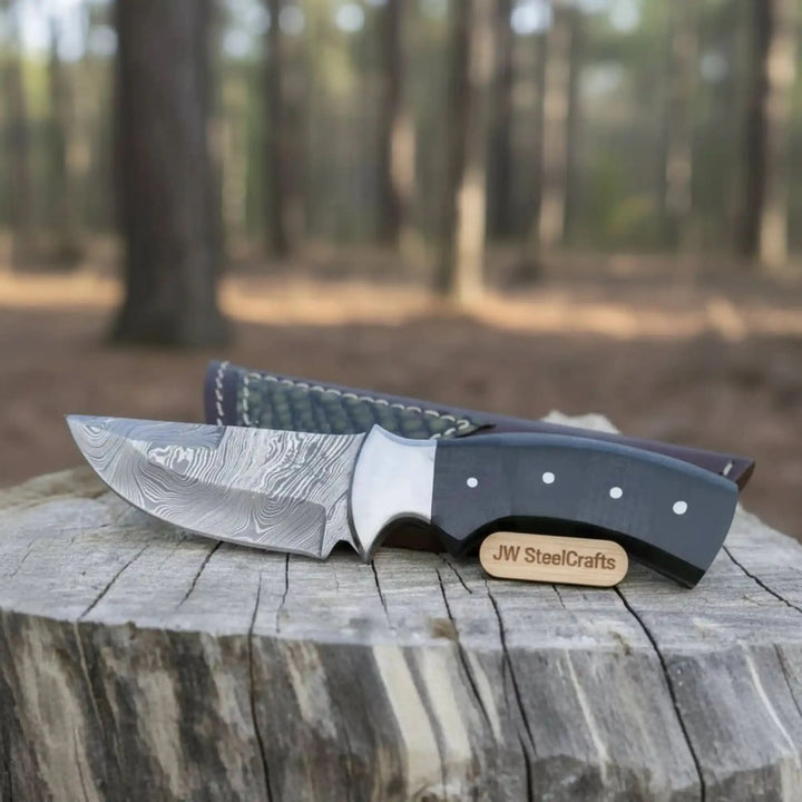Rustic fixed blade Damascus knife with black grip, curved blade, leather sheath, and blurred forest background.