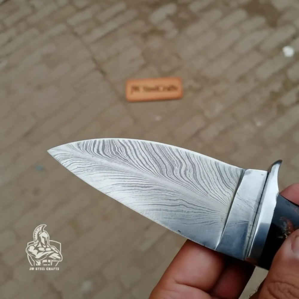 Damascus skinner with black resin handle by JW SteelCrafts, featuring patterned blade, silver guard, and forest table display