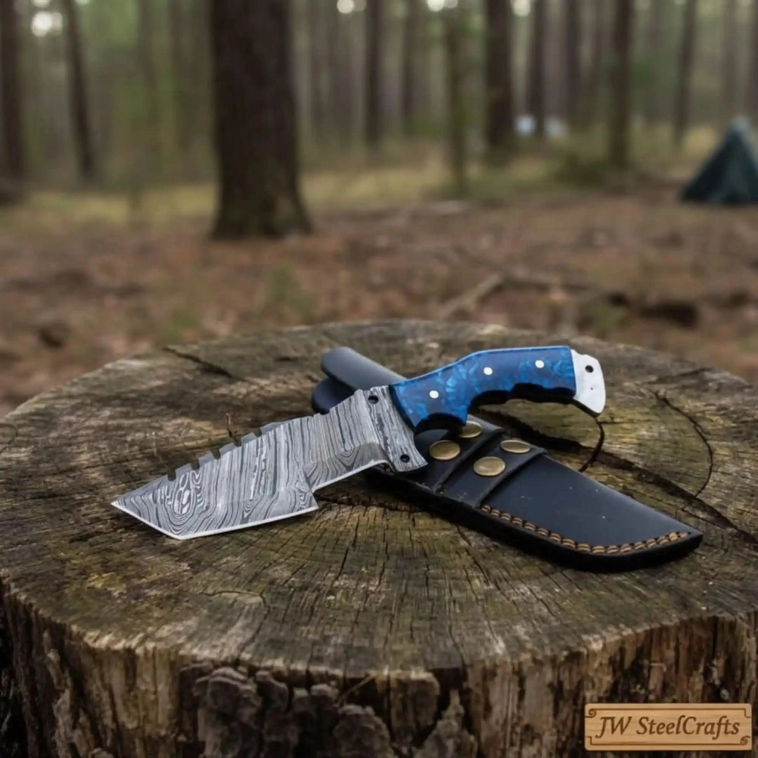 Notched blade custom camp knife with a blue grip, patterned steel blade, and fitted black sheath in a forest setting.