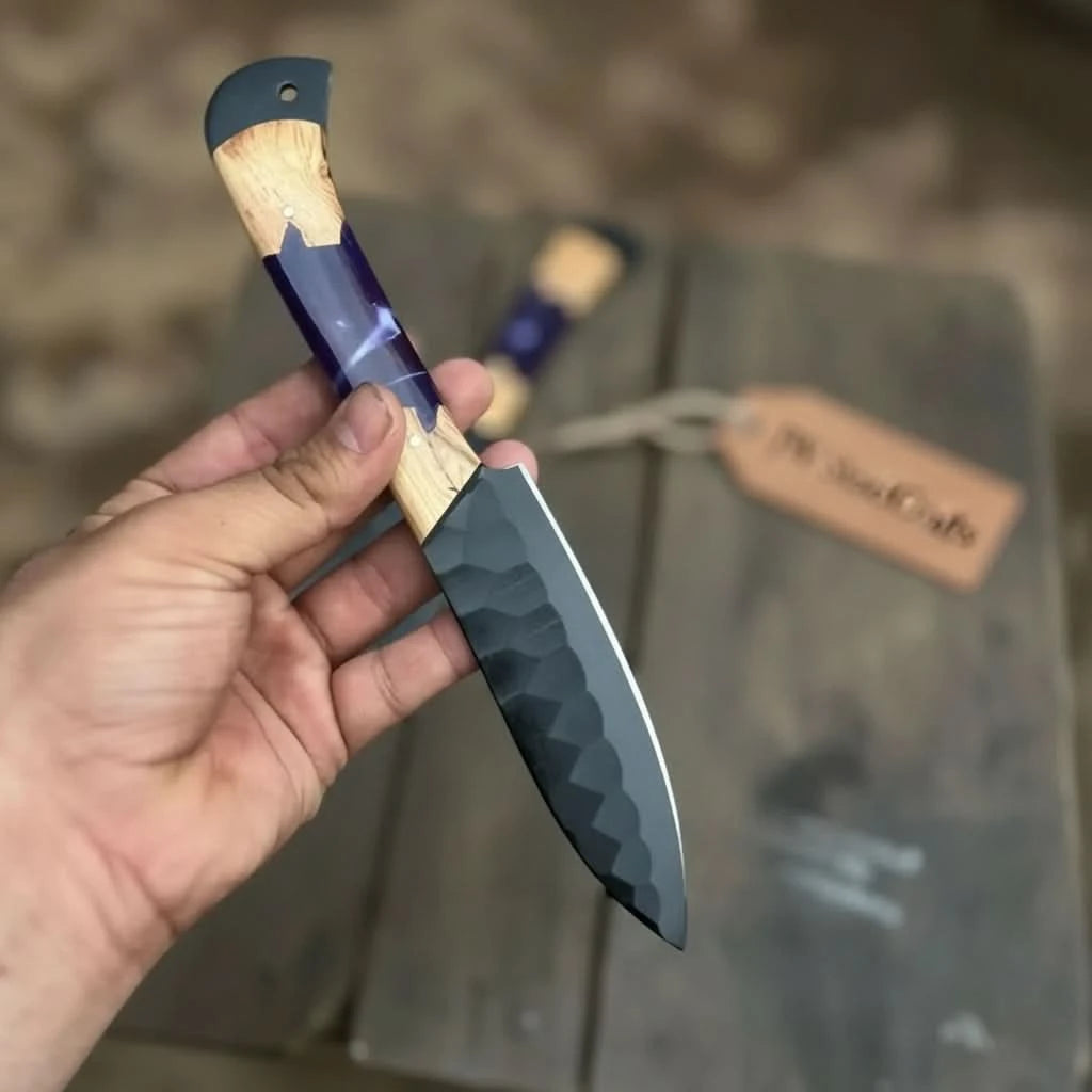 Closeup handheld view of a hand-forged black steel kitchen knife showcasing hammered blade texture and unique purple resin with olive wood combination handle
