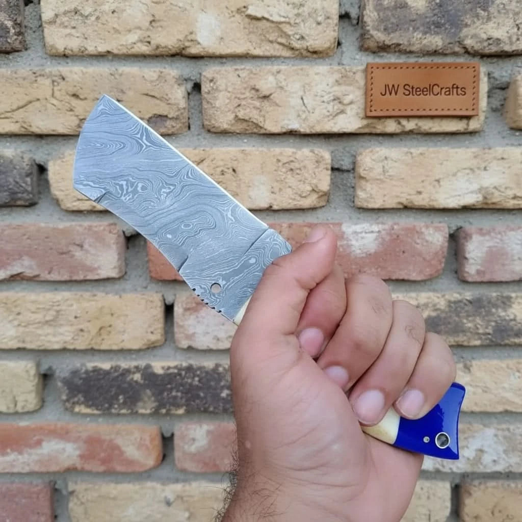 Brick wall handheld Damascus cleaver display showing a broad patterned blade and two-tone handle.