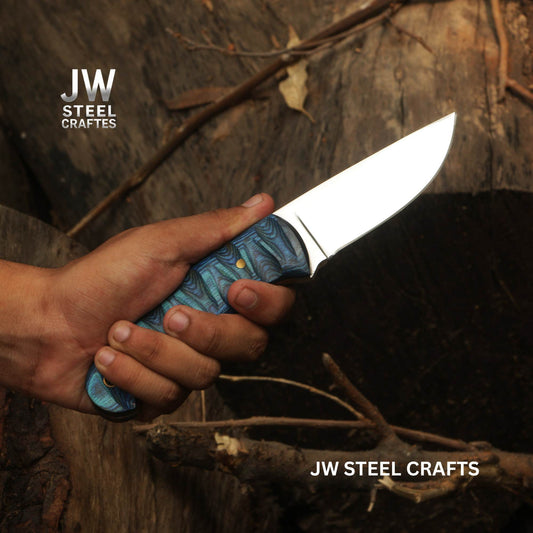 Hand holding a knife with a blue handle against a wooden background, featuring the JW Steel Crafts logo.