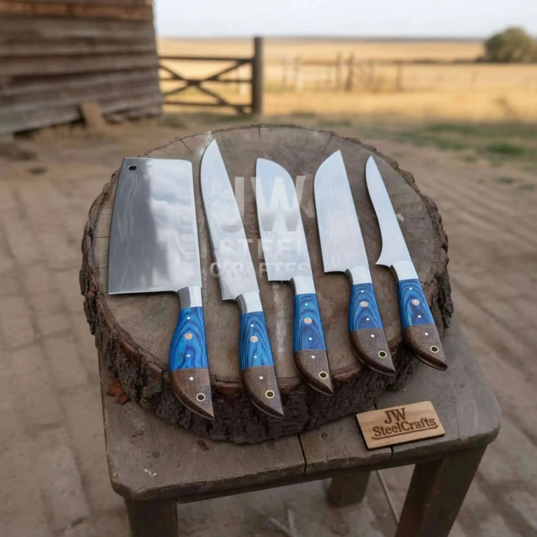 A 5-piece handmade Damascus steel chef knife set with unique blue and brown two-tone wood handles, displayed vertically on a rustic wooden log in an outdoor setting.