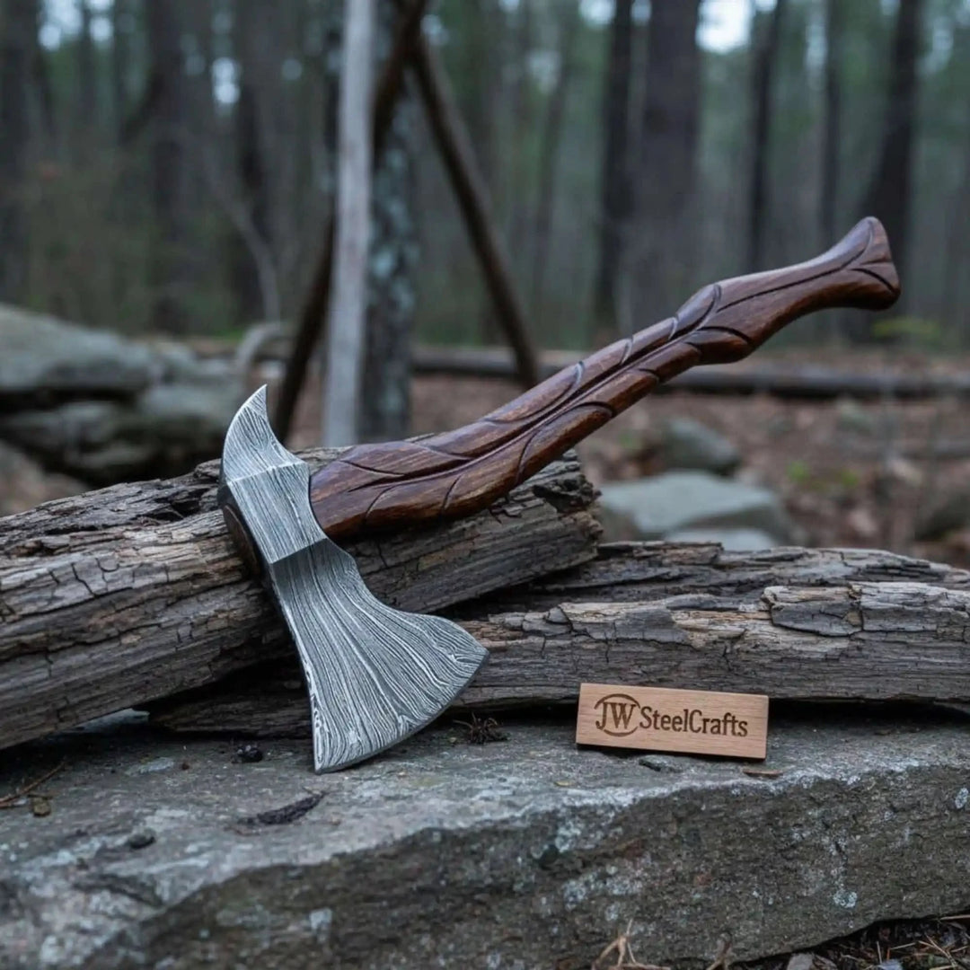     Wilderness Damascus camp axe with curved carved hardwood handle resting on rough timber in forest setting.
