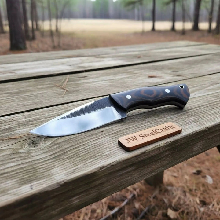 Custom camping knife with wrist strap sheath, black handle, polished blade, and rustic outdoor wood surface.