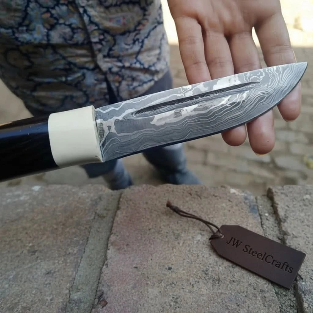 Black handle Damascus knife blade closeup with cream bolster, long polished blade, and visible steel grain.