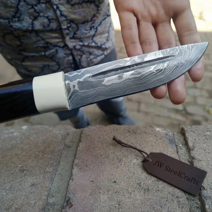 Black handle Damascus knife blade closeup with cream bolster, long polished blade, and visible steel grain.