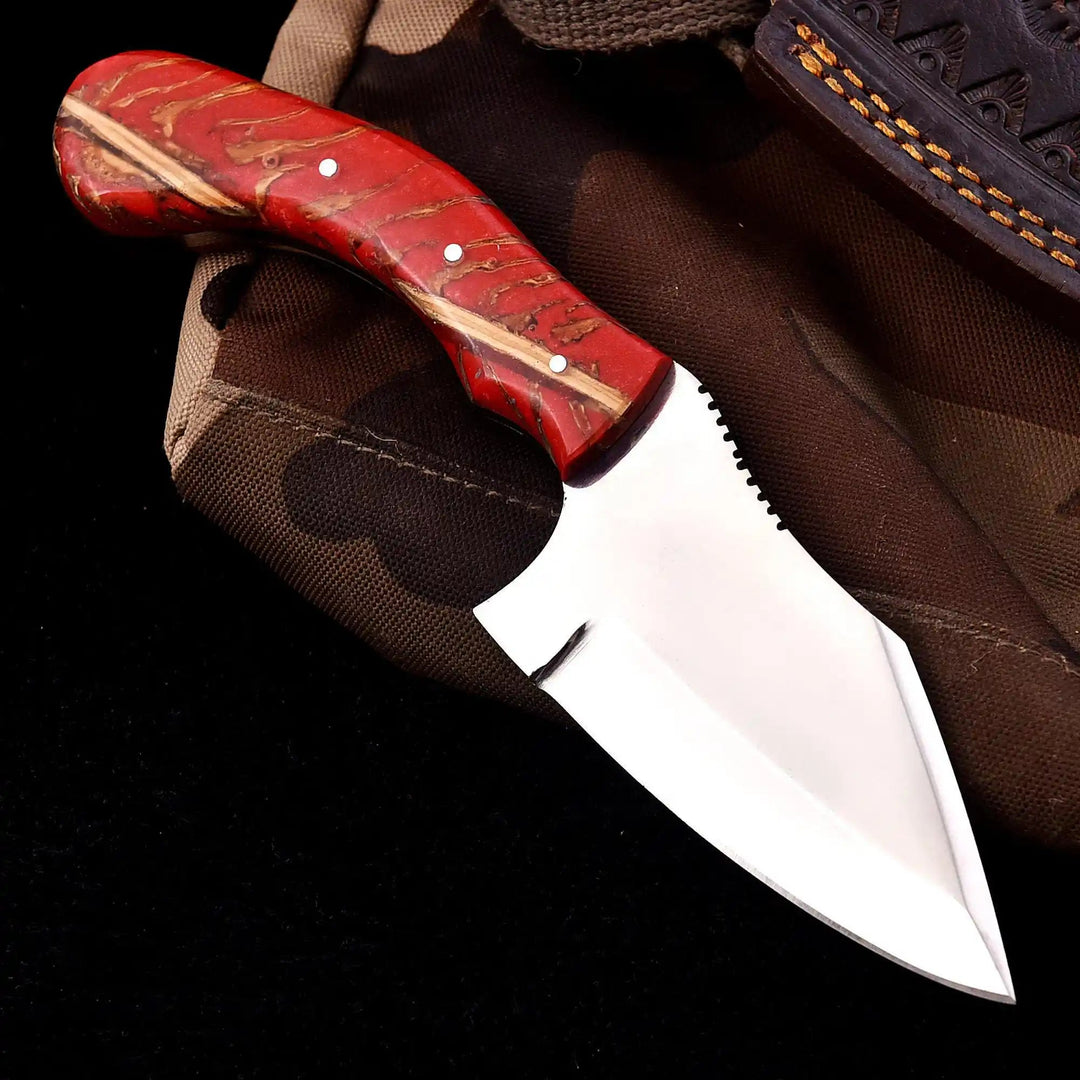 Bright blade handmade knife with a red patterned handle resting beside a stitched leather sheath edge.