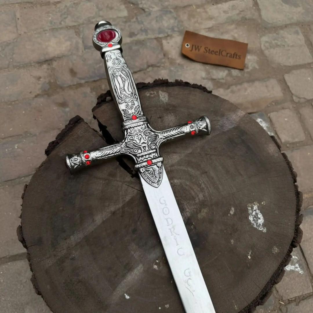 A heritage-style medieval sword with a slim polished silver blade and red gem-studded crossguard, photographed diagonally on a mossy outdoor tree stump.