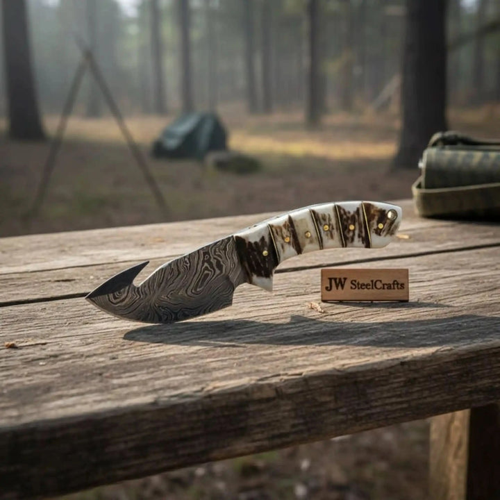 Stag handle Damascus hunting knife with gut hook blade, curved edge, brass pin details, and outdoor forest setting