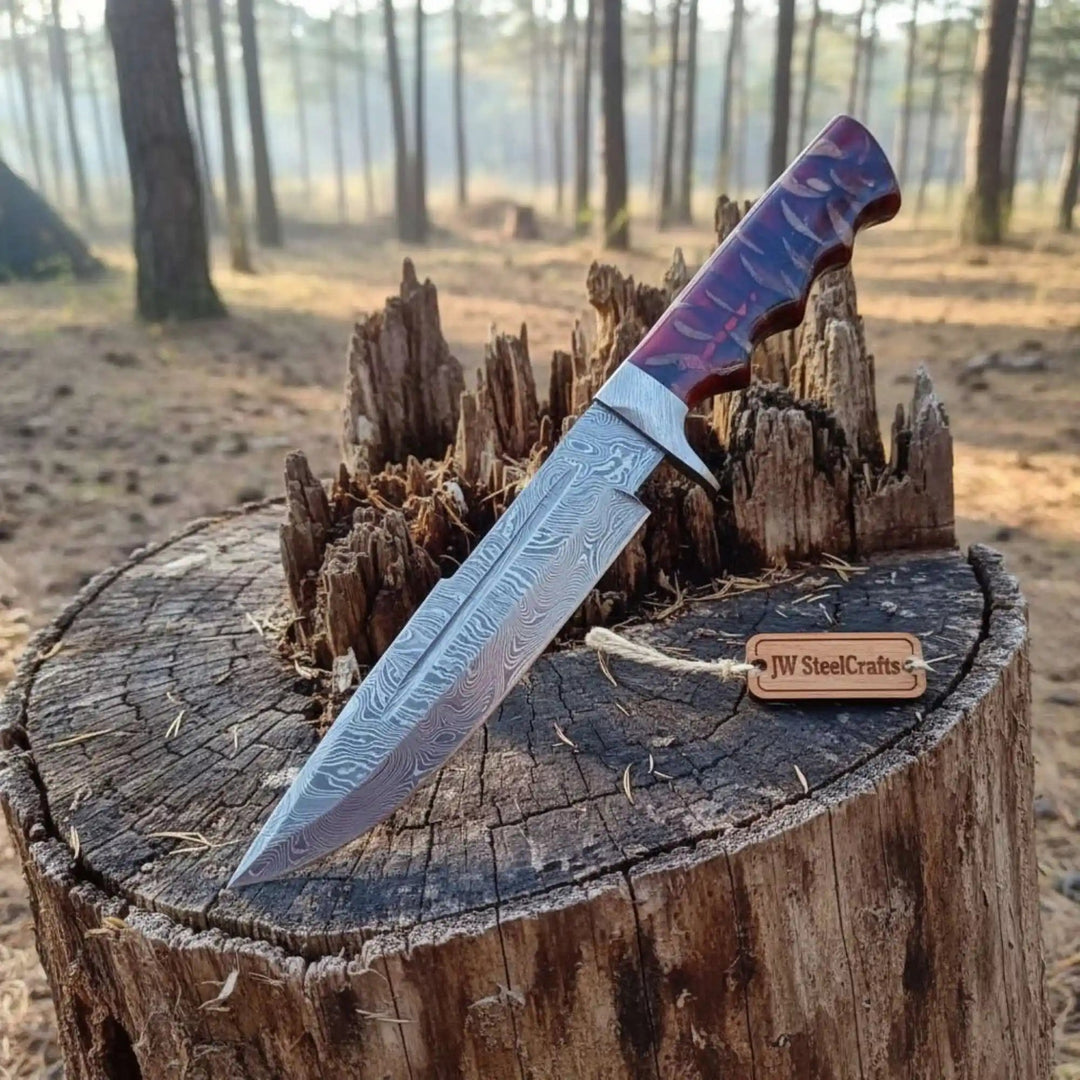 Long Damascus Bowie knife with purple red resin handle, silver guard, and JW SteelCrafts tag on a forest tree stump.