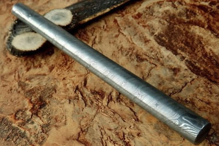 Raw round Damascus steel bar with faint layered pattern placed on textured wooden surface for custom knife making applications