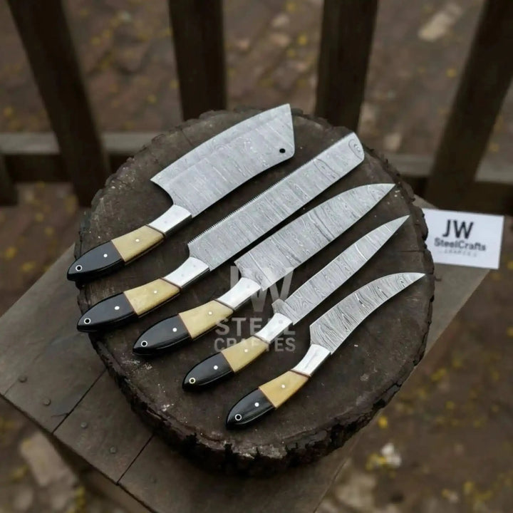 A 5-piece handmade Damascus steel chef knife set with elegant camel bone and black micarta handles, showcased on a rustic wooden log with a scenic blurred outdoor background.