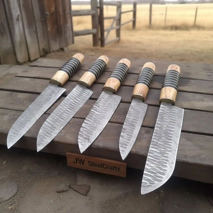 Five handmade Damascus steel chef knives by JW SteelCrafts displayed 
on a rustic wooden stand outdoors, featuring olive wood handles with 
black and silver resin rings and brass bolsters, showing varying blade 
lengths with detailed wave-pattern Damascus steel, brand plaque visible 
in front.