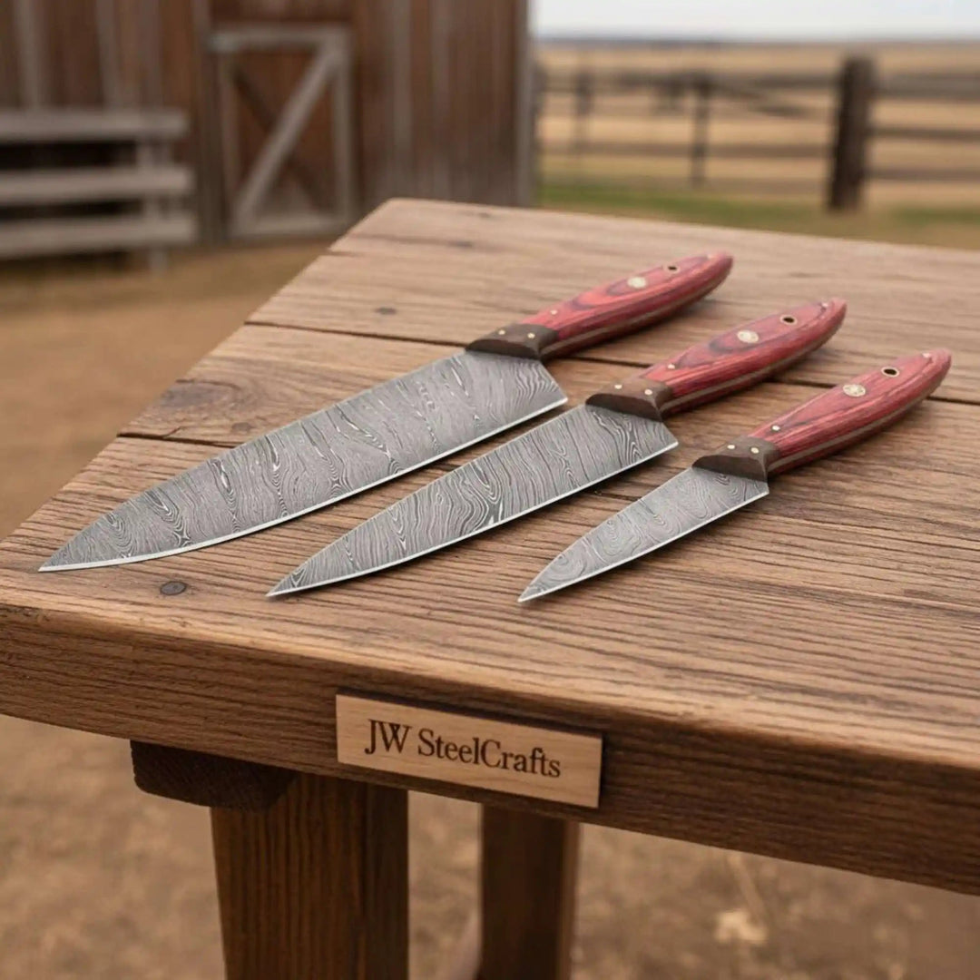 Three handmade Damascus steel kitchen knives by JW SteelCrafts 
arranged on a rustic wooden table, featuring red Pakkawood handles 
with wood bolsters, displayed in graduating sizes outdoors.