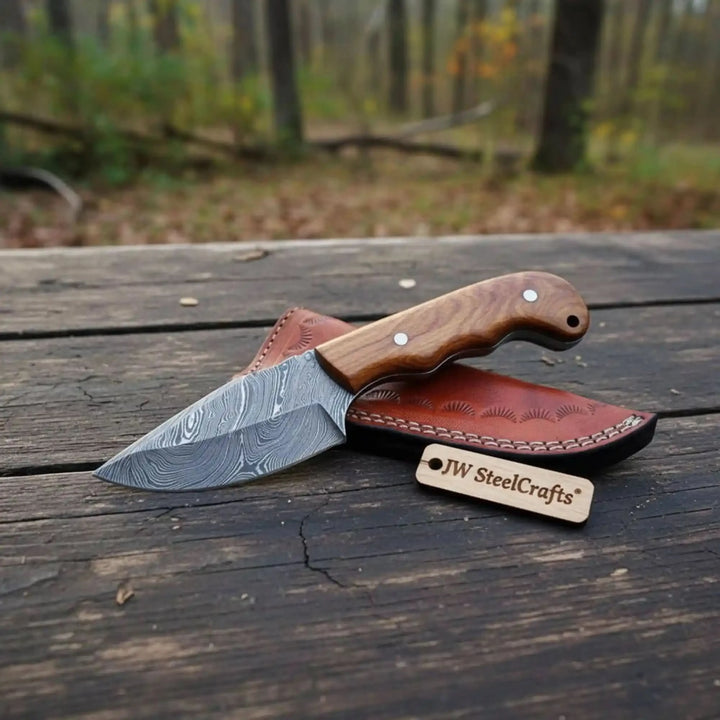 Rustic fixed blade knife with wooden handle, patterned Damascus blade, and brown leather sheath in a woodland setting.