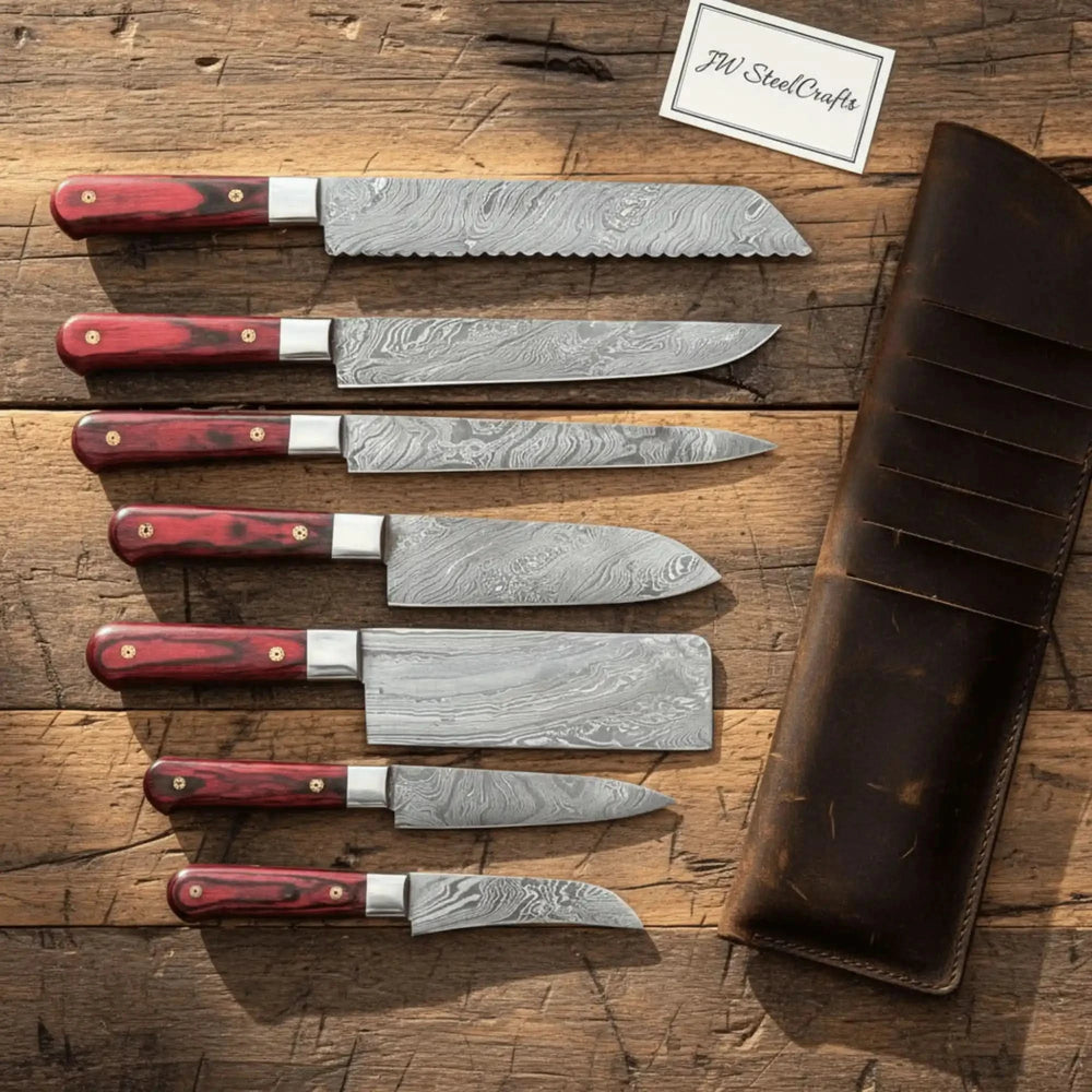 Seven Damascus steel kitchen knives with red wood handles arranged on a rustic wooden table with a leather roll bag.
