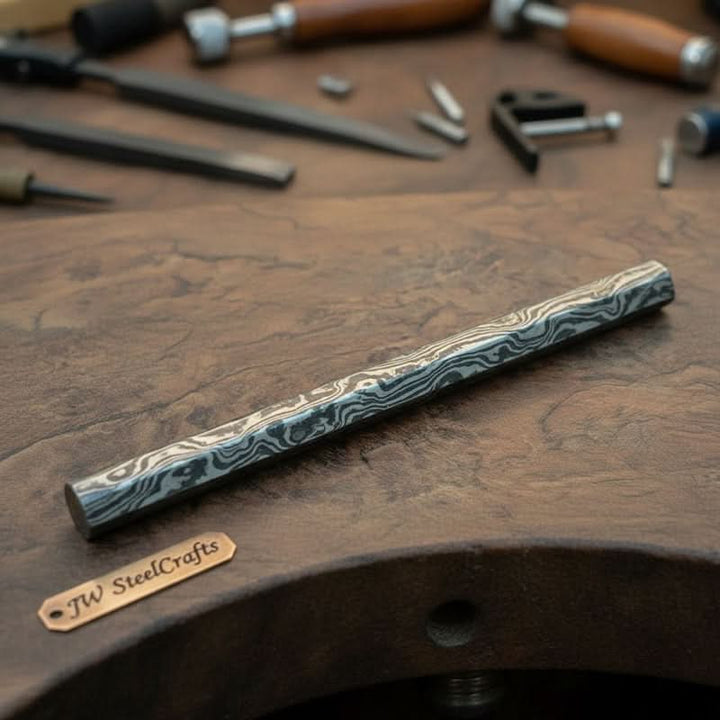 Damascus steel round bar with layered pattern placed on a workshop table beside knife making and metalworking tools