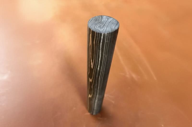 Vertical Damascus steel round bar with layered grain pattern and polished finish displayed on warm background for knife making materials
