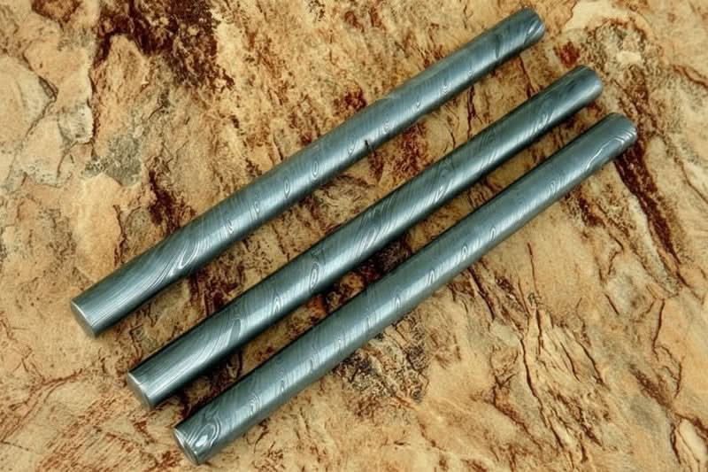 Three round Damascus steel bars with layered grain pattern displayed on textured wooden surface for custom knife making materials