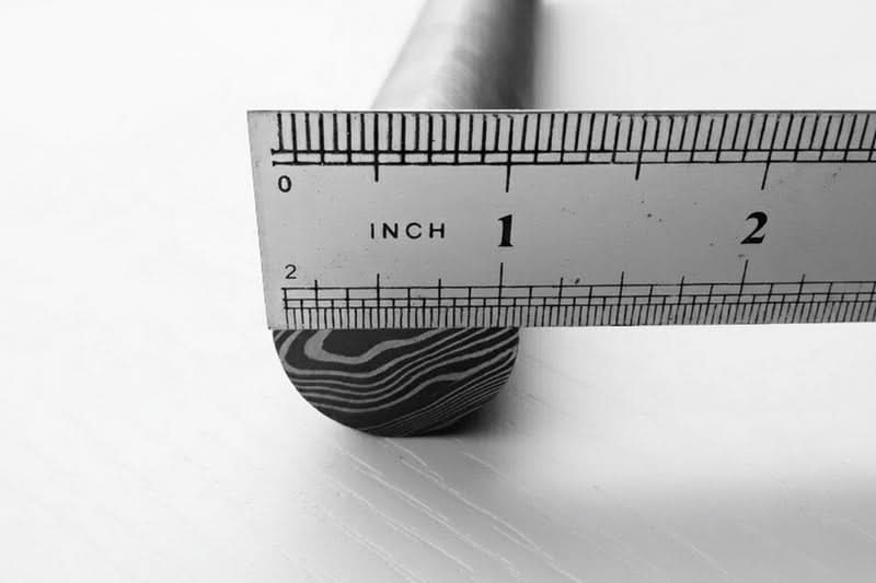 Damascus steel round stock bar with layered end grain pattern positioned under inch ruler for material size reference and measurement