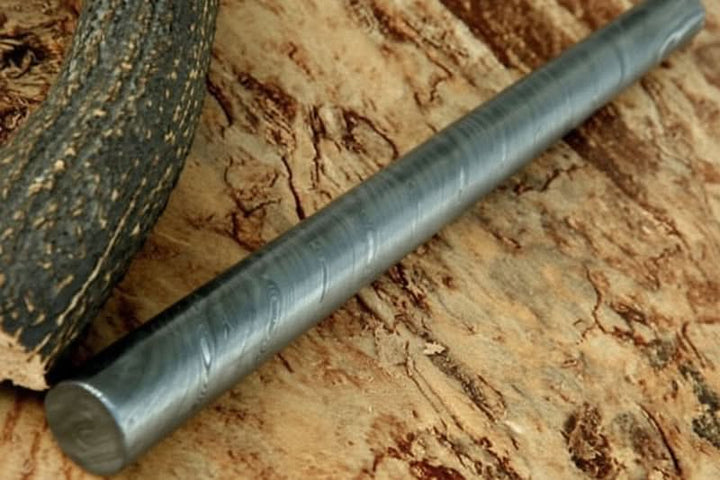 Round Damascus steel stock bar with subtle layered grain pattern displayed on rustic wood surface for handmade knife making materials