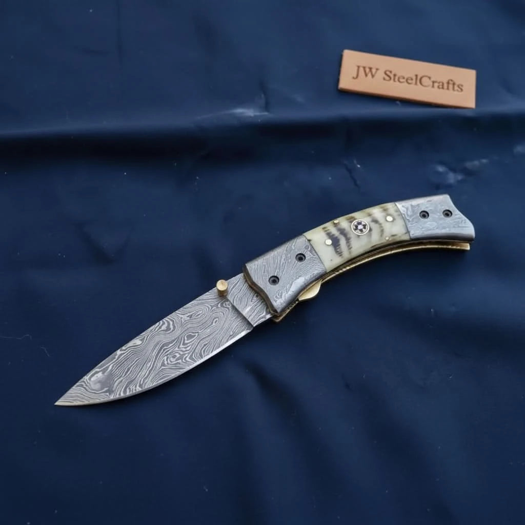 Folding Damascus blade knife with brass thumb stud, bone and metal handle, and dark blue fabric product display.