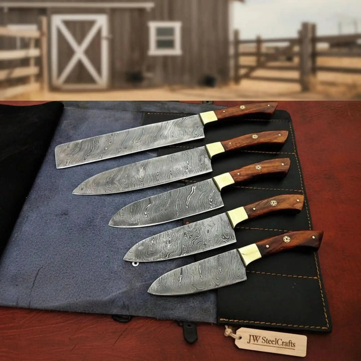 Five Damascus kitchen knives arranged diagonally on dark leather roll with rustic barn background.