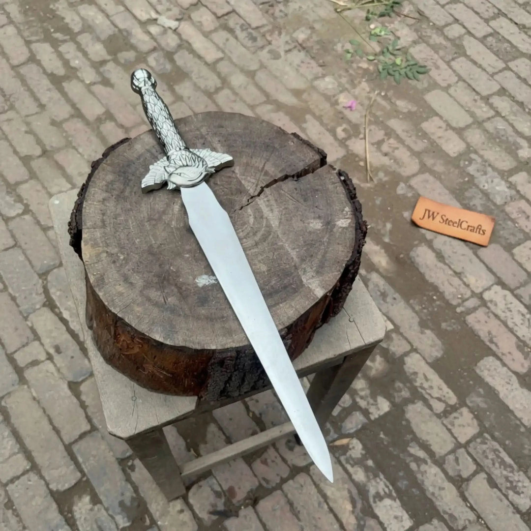 Alongside a weathered tree stump, a medieval-style short sword with a dragon-themed hilt is displayed outdoors on a rustic wooden stand.