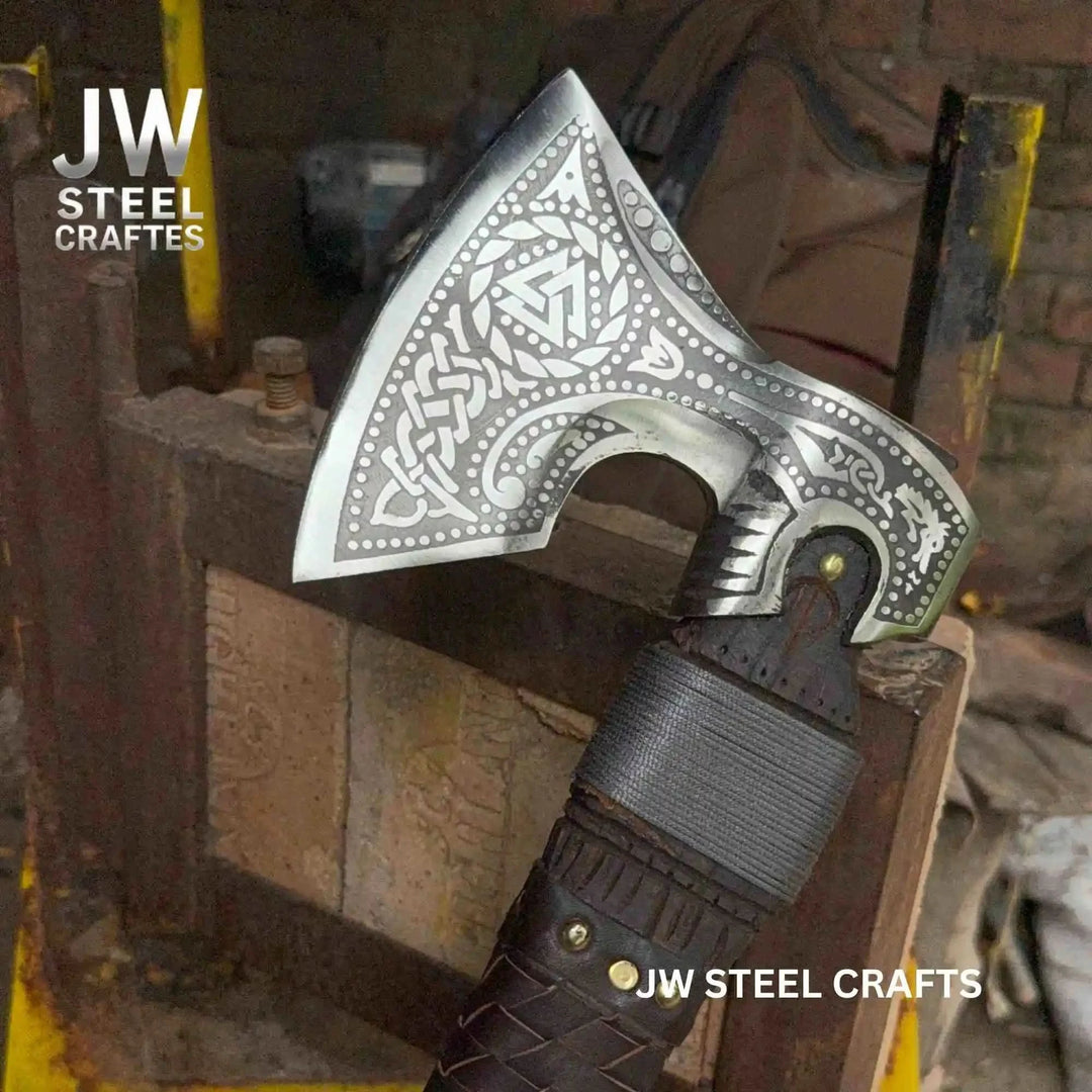 Elegant Nordic war hatchet with ornate blade engravings and dark braided leather handle in workshop surroundings.