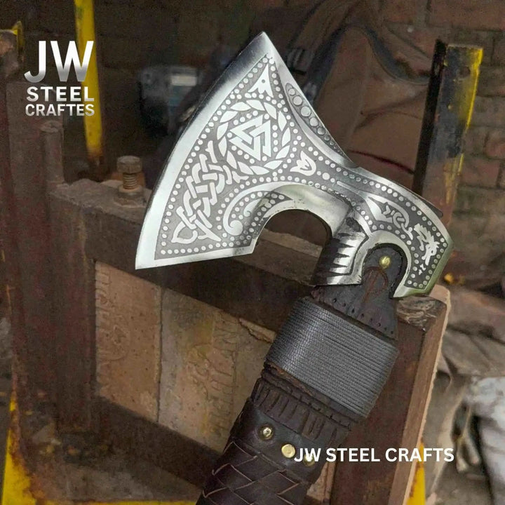 Elegant Nordic war hatchet with ornate blade engravings and dark braided leather handle in workshop surroundings.