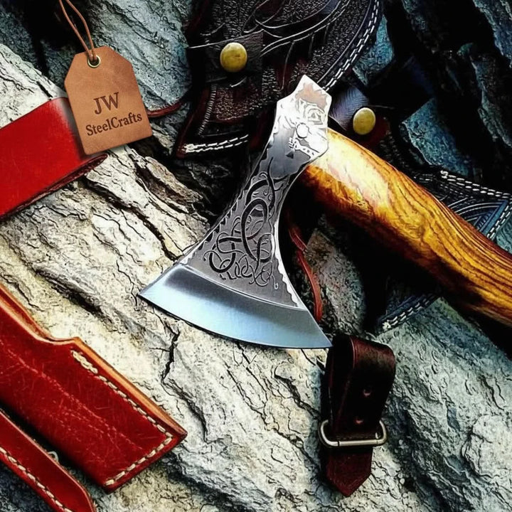     Custom forged camping axe with decorative Nordic engraving and curved wooden grip photographed on a rustic wooden table.