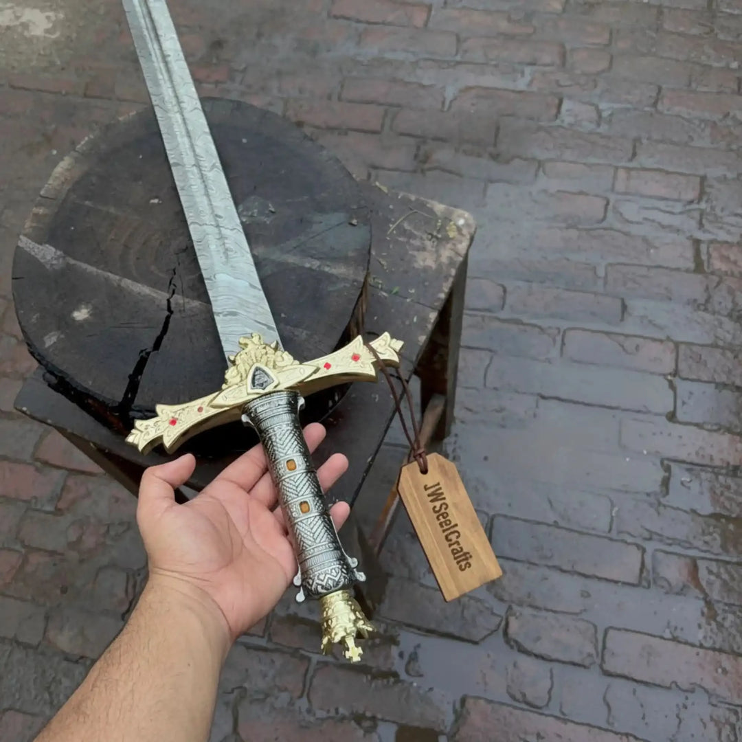 Close-up product display of a JW SteelCrafts Damascus longsword showing the intricate gold hilt details, red gem accents, and swirling Damascus blade pattern.