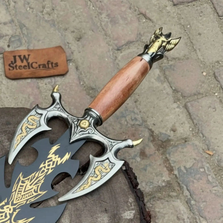 Close-up of an ornate fantasy battle axe featuring gold dragon engravings on a silver guard, a smooth wood grip, and a detailed winged dragon pommel, resting on cobblestones.