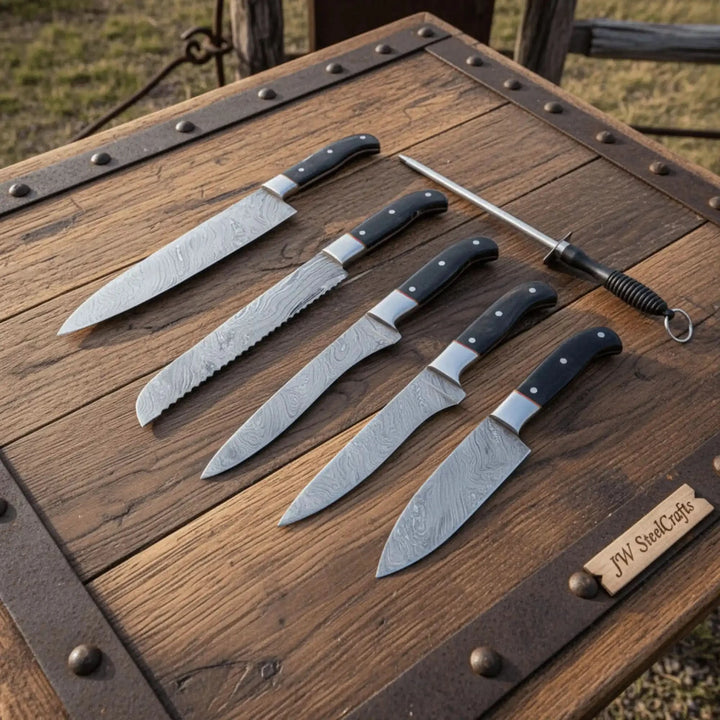 Five handcrafted Damascus steel kitchen knives with black ergonomic handles displayed beside honing steel on rustic wooden outdoor table.