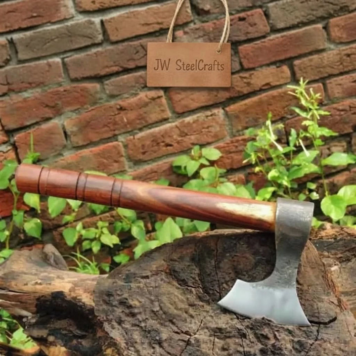 carbon steel camping axe with polished wooden handle displayed on a rustic tree stump against a brick wall background 