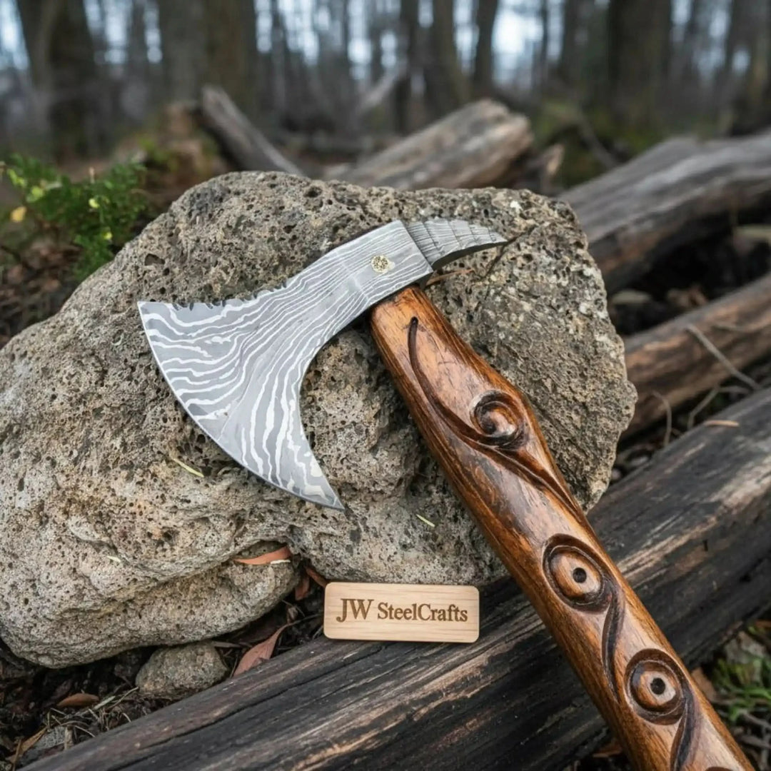 Damascus bearded axe with engraved steel blade and carved wooden handle displayed on natural stone in a forest setting 