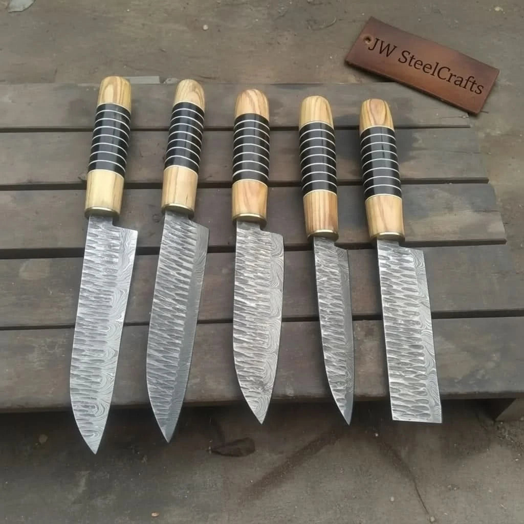 Top-down view of five handmade Damascus steel kitchen knives 
arranged on a dark wooden surface, each with olive wood handles 
wrapped in black and silver resin rings, displaying varied blade 
shapes including chef, santoku, and utility styles with 
wave-patterned Damascus steel.