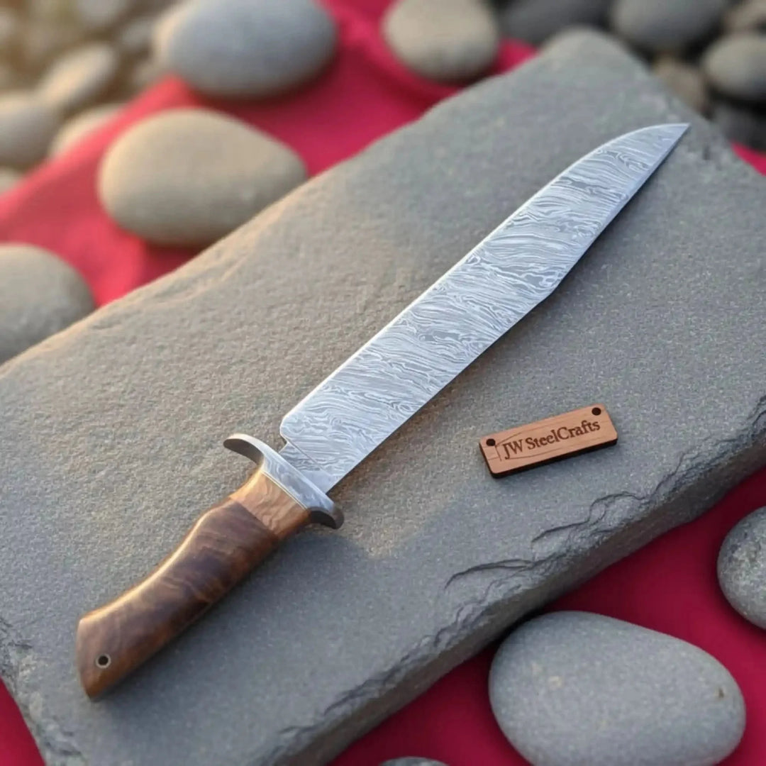 Handmade Damascus Steel Fixed Blade Hunting Knife With A Wooden Handle And 'JW SteelCrafts' Label On A Stone Surface With Pebbles In The Background
