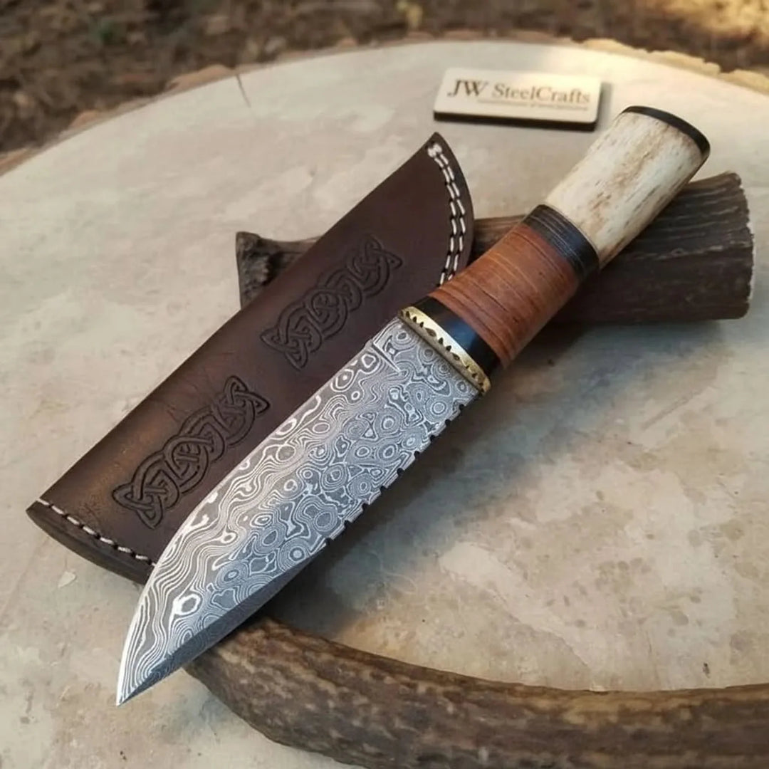 Handmade damascus steel hunting knife with a wooden handle and leather sheath displayed on a rustic surface.