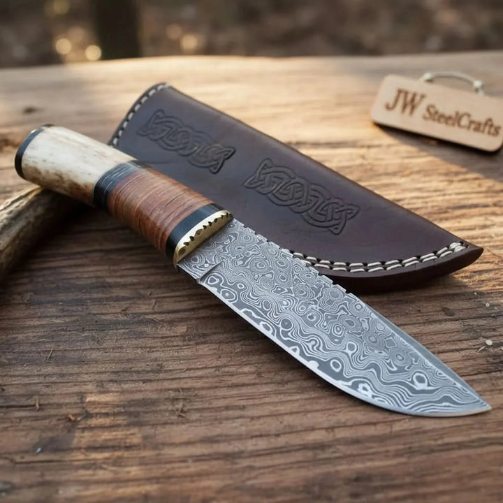 Handmade damascus steel hunting knife with a wooden handle and leather sheath displayed on a wooden surface.
