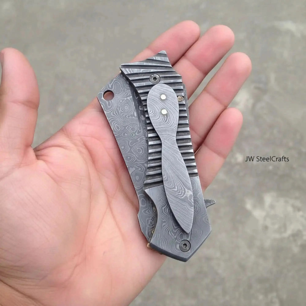 Handmade EDC Damascus Flipper Pocket Knife Folding Knife With Textured Blade Held In A Hand Against A Neutral Background.
