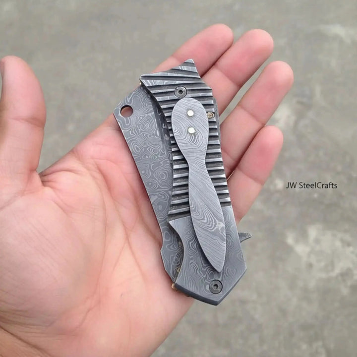 Handmade EDC Damascus Flipper Pocket Knife Folding Knife With Textured Blade Held In A Hand Against A Neutral Background.
