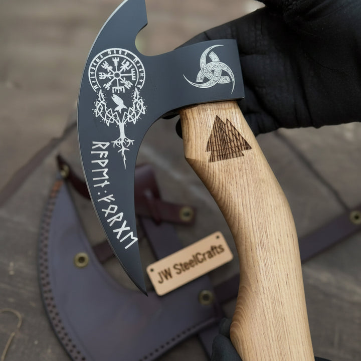 Artisan-forged bushcraft axe with decorative black steel blade and stitched leather sheath photographed in a forest setting.