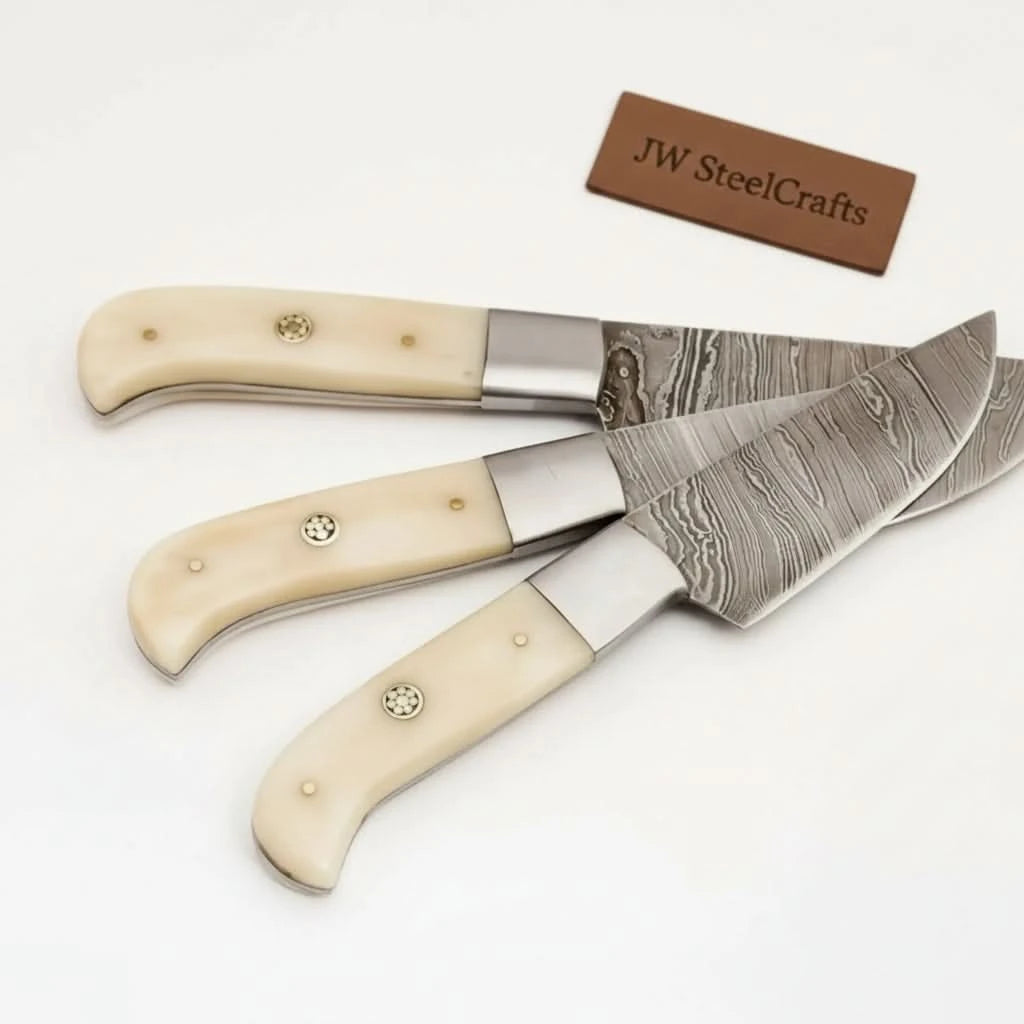 Three Damascus steel kitchen knives arranged horizontally on white background with smooth ivory colored ergonomic handles.