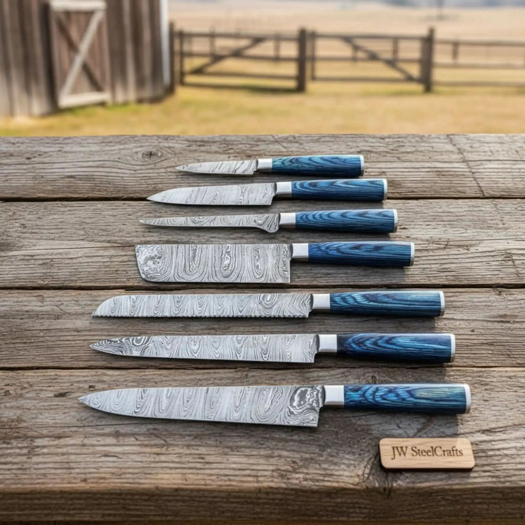 Damascus steel chef knives arranged in size order on weathered wood with outdoor countryside background.