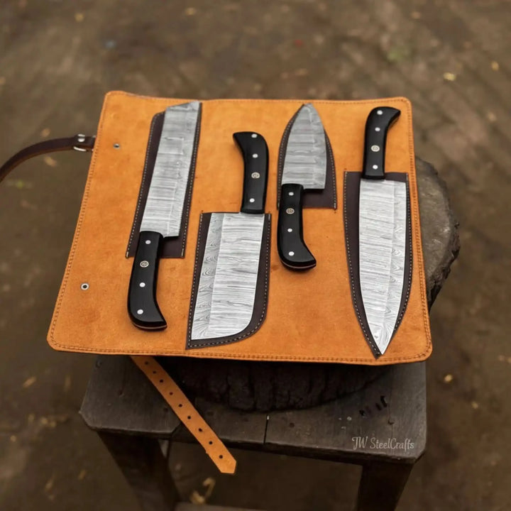 Set of four culinary knives with ebony handles covered in individual hand-stitched dark leather blade guards, arranged on an unrolled caramel suede knife roll case
